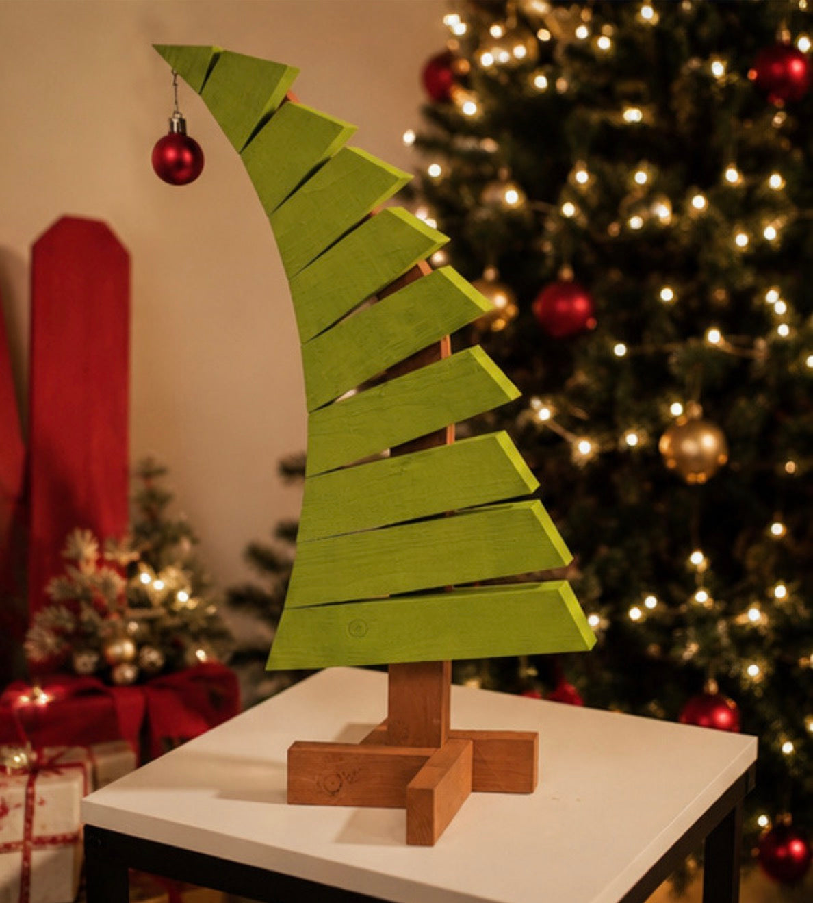 Green wooden Christmas tree with red ornament displayed on a white tabletop beside a decorated Christmas tree